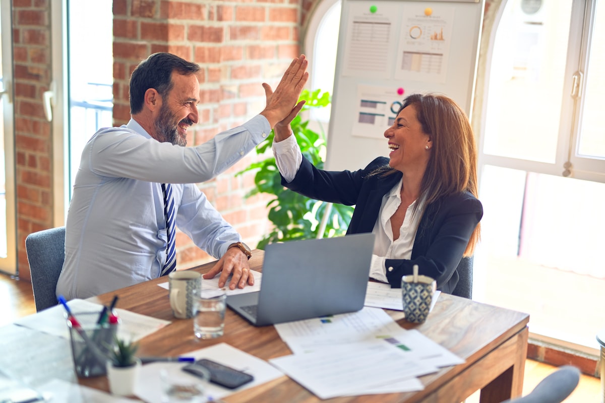 Two colleagues celebrating with a high five at a desk with a laptop and printed charts
