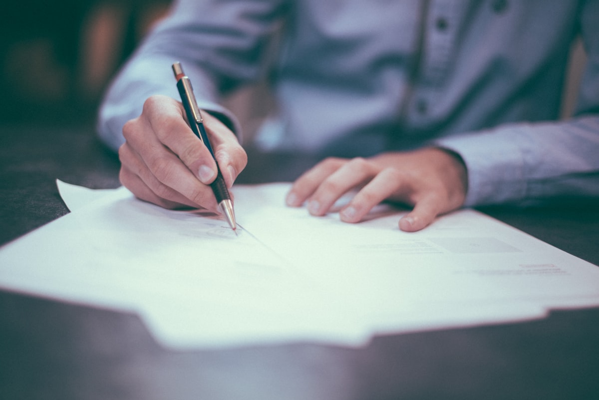 Person in a light blue shirt writing with a pencil on printed documents at a desk
