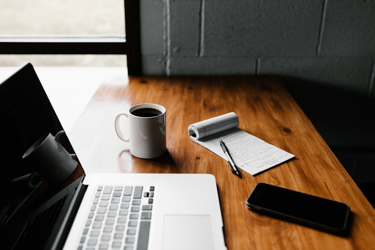 Open laptop, a mug of black coffee and a notepad with handwritten notes on a wooden desk
