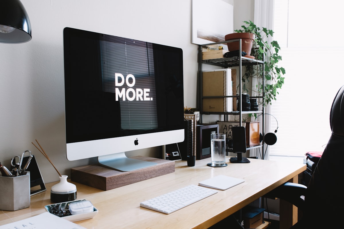 Tidy wooden desk with an iMac showing the words Do More, a keyboard, and a shelf with notebooks and a plant