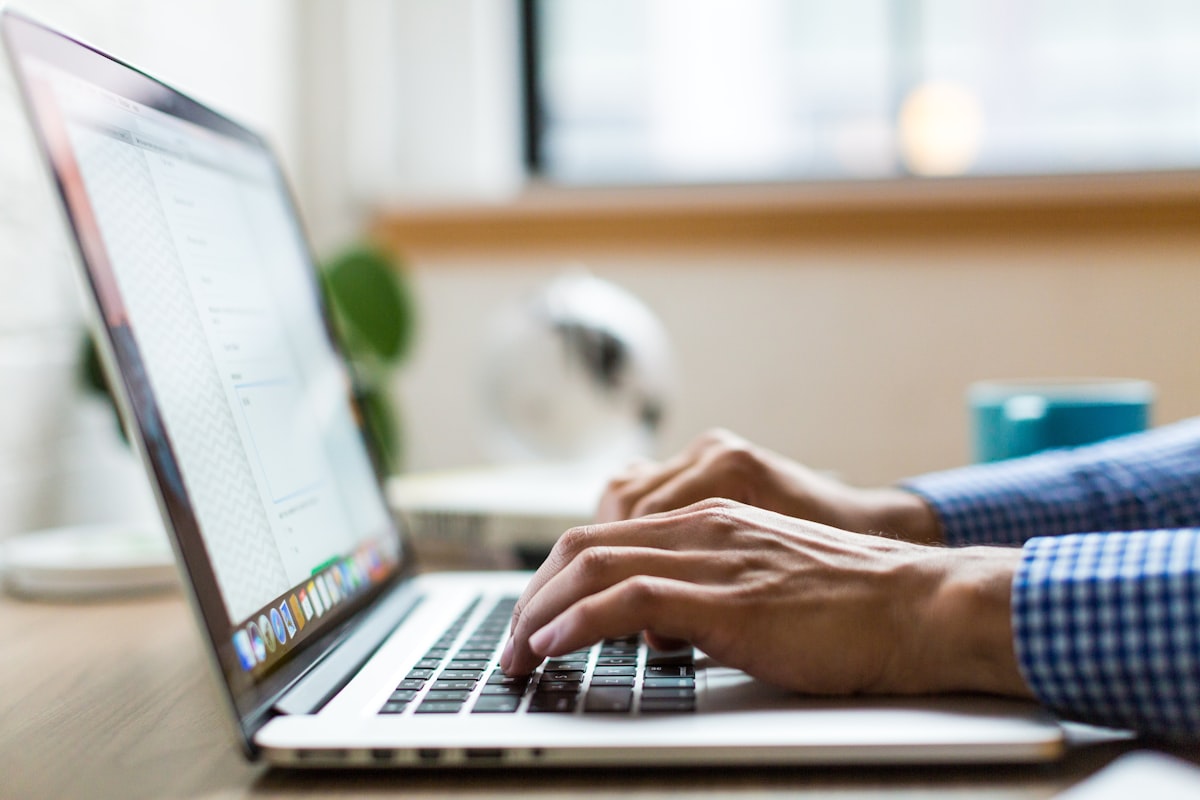 Close-up of hands typing on a laptop keyboard at a bright desk with a blue mug in the background