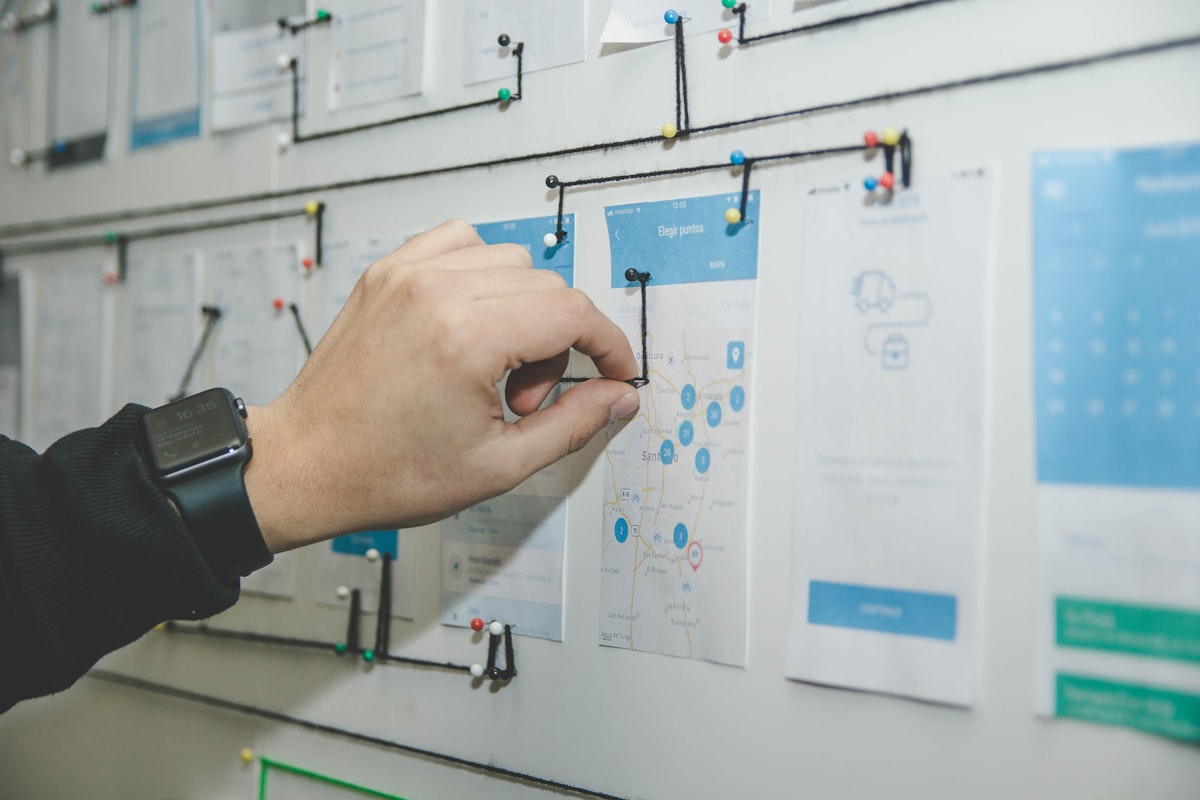 Hand pinning printed mobile app screen designs to a wall, connected with black string to map the user flow