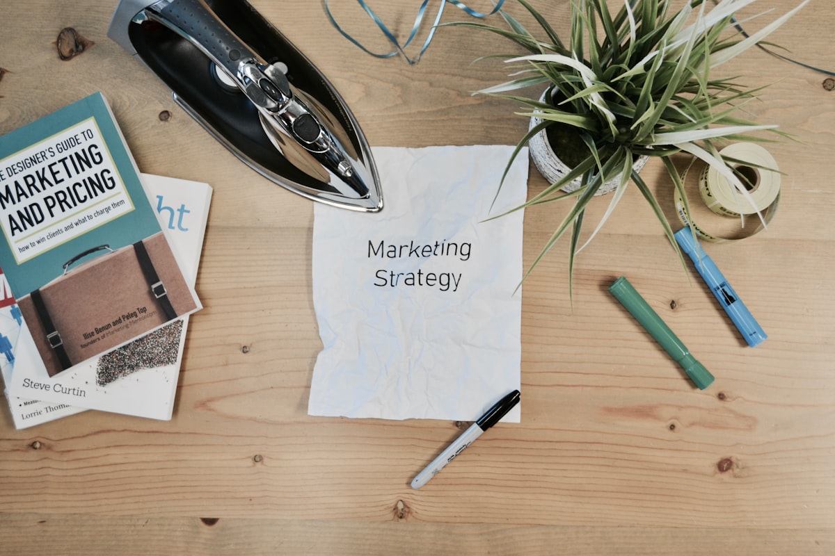 Flat-lay of a wooden desk with a note reading Marketing Strategy, coloured markers, a plant and marketing books