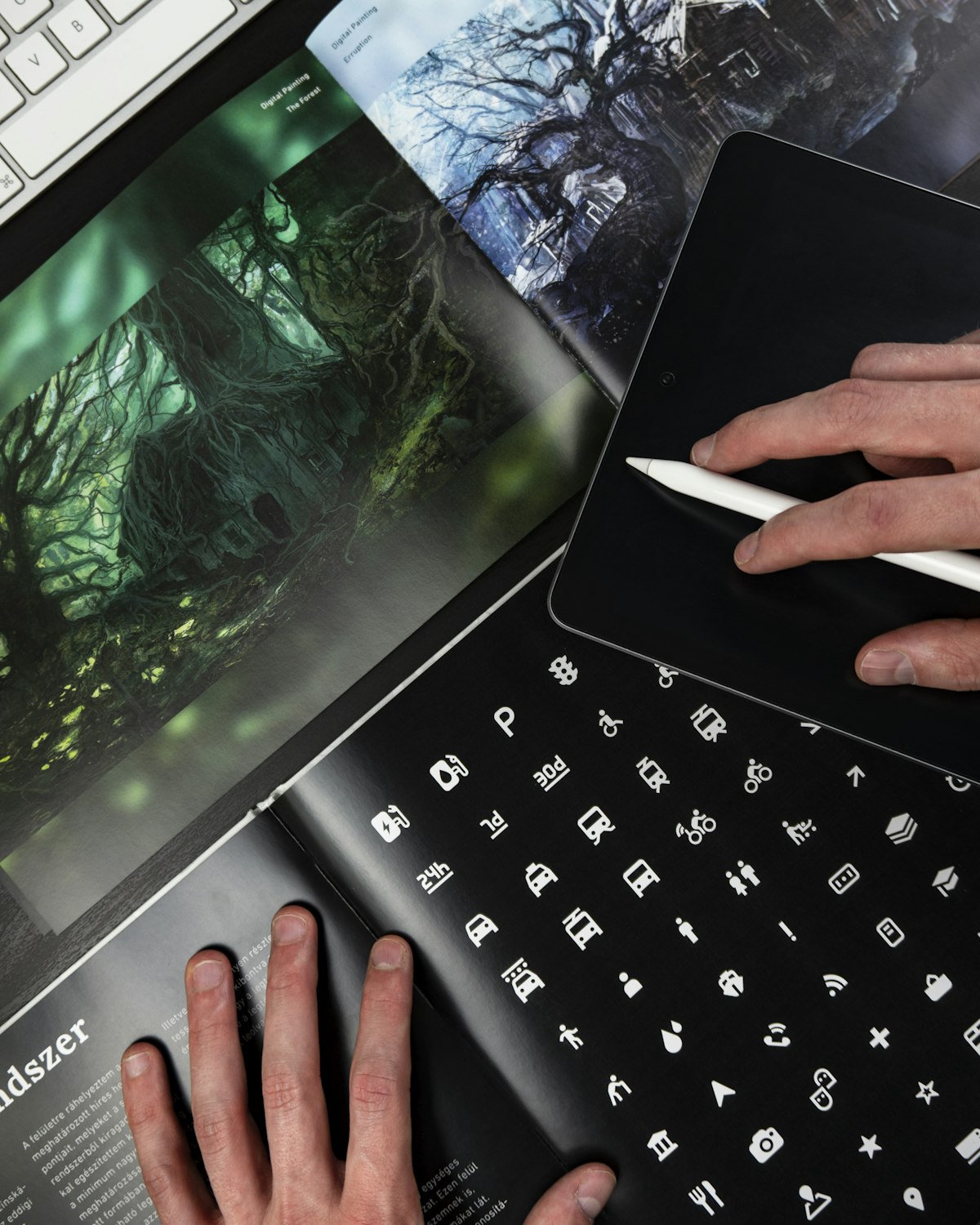 A designer with a tablet and stylus beside a magazine page full of small icon designs
